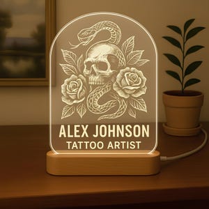 Puede incluir: Un letrero acrílico iluminado con un diseño de calavera, serpiente y rosas. El letrero está personalizado con el nombre "ALEX JOHNSON" y el texto "TATTOO ARTIST". La base es de madera clara.