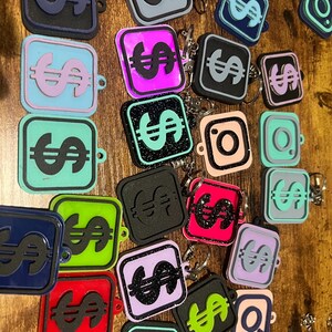 Könnte beinhalten: Eine Sammlung bunter quadratischer Schlüsselanhänger mit einem Dollarzeichen oder dem Instagram-Logo. Die Schlüsselanhänger sind aus verschiedenen Materialien hergestellt, darunter Glitzer, glatter Kunststoff und strukturierter Kunststoff.