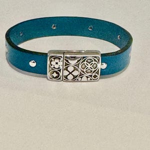 May include: A teal leather bracelet with a silver magnetic clasp featuring a geometric design.