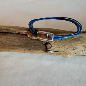 Puede incluir: Un brazalete de cuero azul con un cierre magnético plateado. El brazalete está hecho de múltiples hebras de cuero.