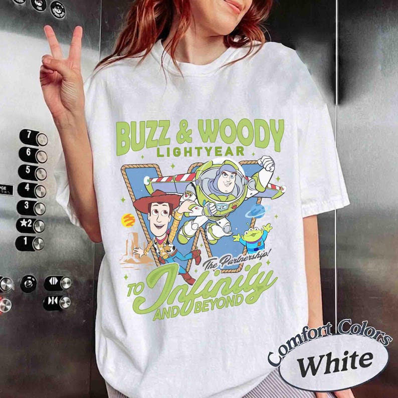May include: A white short-sleeve t-shirt featuring a graphic of Buzz Lightyear, Woody, and a green alien. The text reads BUZZ & WOODY LIGHTYEAR and TO INFINITY AND BEYOND. The design includes a blue triangular background and yellow stars.