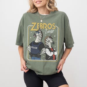 May include: Olive green t-shirt featuring a cartoon graphic of two zebras. One zebra is wearing a police vest, and the other is in a letterman jacket. The text "ZEBROS" and "Zebras" is above the zebras in a yellow frame.