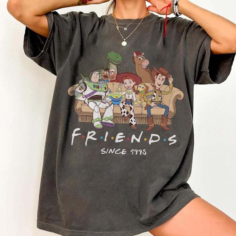 May include: A charcoal gray short-sleeve t-shirt featuring a graphic of Toy Story characters sitting on a couch. The text below the characters reads "F R I E N D S" with colorful dots between letters and the text "SINCE 1995" underneath.