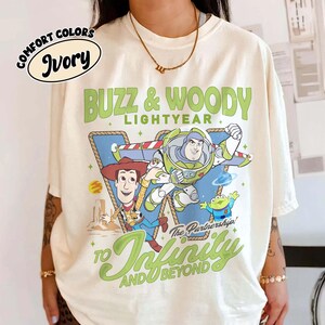 May include: An ivory t-shirt featuring a graphic of Buzz Lightyear, Woody, and an alien from Toy Story. The text reads BUZZ & WOODY LIGHTYEAR, The Partnership!, and TO INFINITY AND BEYOND. A small oval label in the top left corner says COMFORT COLORS IVORY.