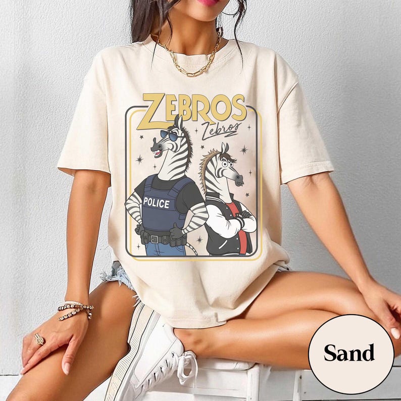 May include: Sand-colored t-shirt featuring a cartoon graphic of two zebras. One zebra wears sunglasses and a police vest, the other a jacket. The text "ZEBROS" is above the zebras. The shirt is short-sleeved.