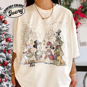 May include: An ivory t-shirt featuring a festive illustration of Disney characters including Mickey Mouse, Minnie Mouse, Donald Duck, Goofy, Pluto, and Chip 'n Dale singing carols in a snowy village. The text COMFORT COLORS IVORY is printed in the corner.