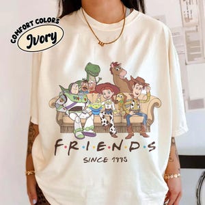 May include: An ivory short-sleeve t-shirt featuring a graphic print of Toy Story characters sitting on a couch with the text "FRIENDS SINCE 1995" below. The characters include Buzz Lightyear, Rex, Mr. Potato Head, Alien, Jessie, Slinky Dog, Bullseye, and Woody.