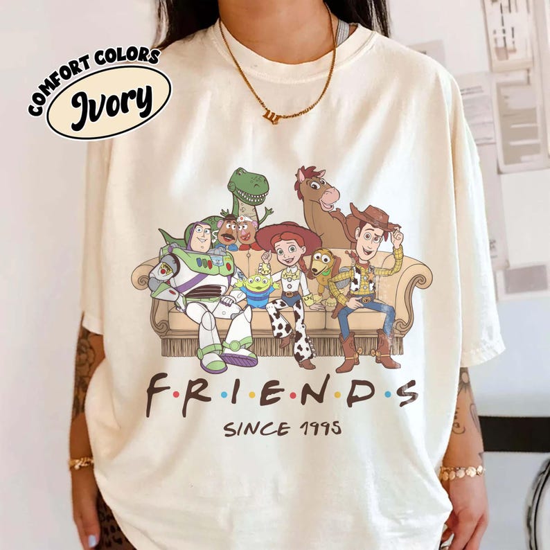 May include: An ivory short-sleeve t-shirt featuring a graphic print of Toy Story characters sitting on a couch with the text "FRIENDS SINCE 1995" below. The characters include Buzz Lightyear, Rex, Mr. Potato Head, Alien, Jessie, Slinky Dog, Bullseye, and Woody.