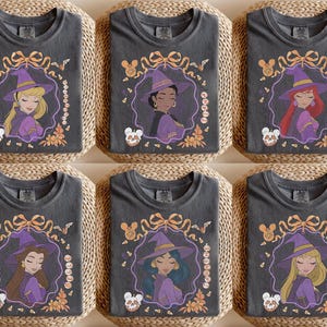 May include: A charcoal gray t-shirt featuring a graphic of a princess wearing a purple witch hat and cape. The design includes a decorative frame with a bow, bats, candy corn, and a skull. Each shirt displays a different princess name in text.