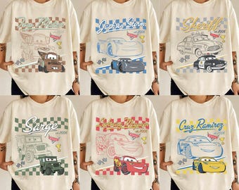 May include: Six cream-colored t-shirts featuring cartoon car designs. Each shirt displays a different character: Tow Mater, Jackson Storm, Sheriff, Sarge, Lightning McQueen, and Cruz Ramirez. The designs include the character's name and the year 2006.