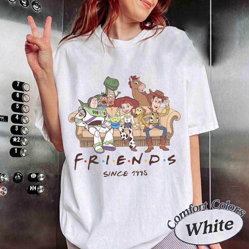 May include: A white short-sleeve t-shirt featuring a graphic of Toy Story characters sitting on a couch with the text "FRIENDS" and "SINCE 1995" printed below. The characters include Buzz Lightyear, Rex, Jessie, Bullseye, and Woody.