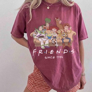 May include: A maroon short-sleeve t-shirt featuring a graphic of Toy Story characters sitting on a couch. The text reads FRIENDS SINCE 1995 in white lettering. The characters include Buzz Lightyear, Rex, Jessie, Slinky Dog, and Woody.
