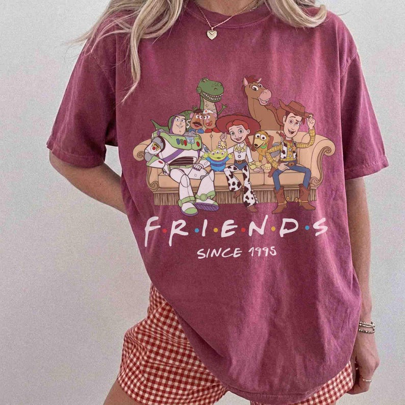 May include: A maroon short-sleeve t-shirt featuring a graphic of Toy Story characters sitting on a couch. The text reads FRIENDS SINCE 1995 in white lettering. The characters include Buzz Lightyear, Rex, Jessie, Slinky Dog, and Woody.