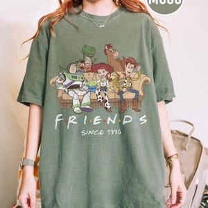 May include: A moss green short-sleeve t-shirt featuring a graphic print of various animated characters sitting on a couch. Below the characters, the text reads FRIENDS SINCE 1995, with colorful dots between the letters of the word FRIENDS.