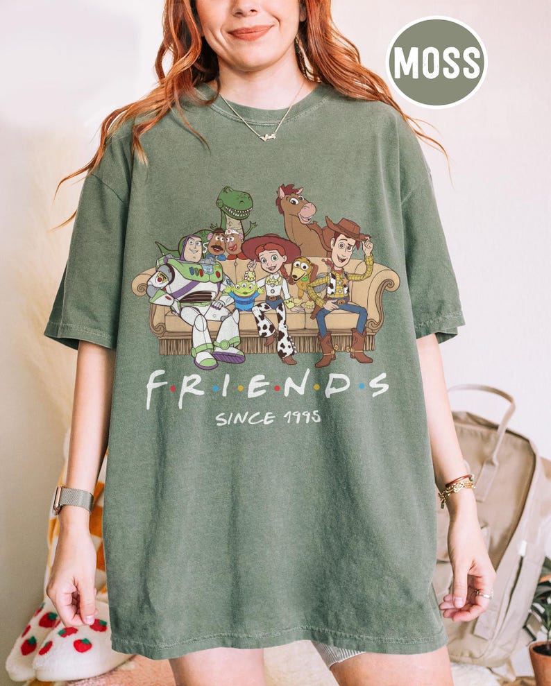 May include: A moss green short-sleeve t-shirt featuring a graphic print of various animated characters sitting on a couch. Below the characters, the text reads FRIENDS SINCE 1995, with colorful dots between the letters of the word FRIENDS.