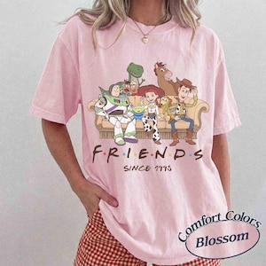 May include: A light pink short-sleeve t-shirt featuring a graphic of Toy Story characters sitting on a couch with the text "F.R.I.E.N.D.S SINCE 1995" printed below. The characters include Buzz Lightyear, Rex, Mr. Potato Head, Jessie, Bullseye, and Woody.