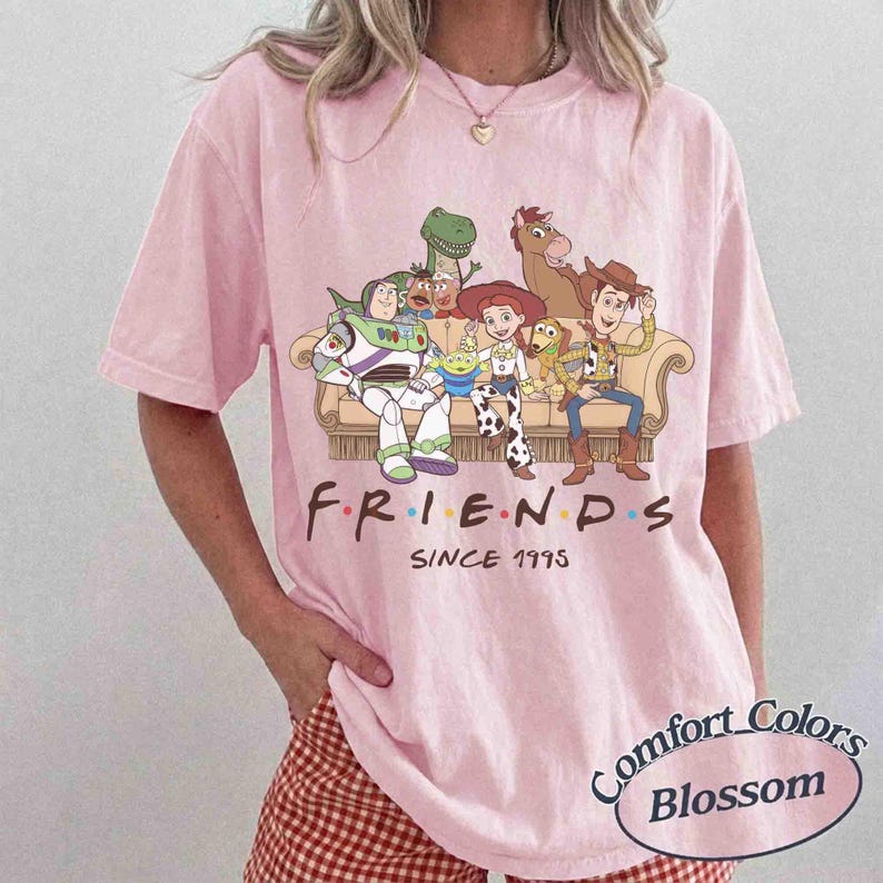 May include: A light pink short-sleeve t-shirt featuring a graphic of Toy Story characters sitting on a couch with the text "F.R.I.E.N.D.S SINCE 1995" printed below. The characters include Buzz Lightyear, Rex, Mr. Potato Head, Jessie, Bullseye, and Woody.