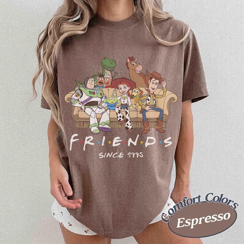 May include: A brown short-sleeve t-shirt featuring a graphic of Toy Story characters sitting on a couch with the text "FRIENDS SINCE 1995" printed below. The characters include Buzz Lightyear, Rex, Mr. Potato Head, Jessie, Slinky Dog, and Woody.