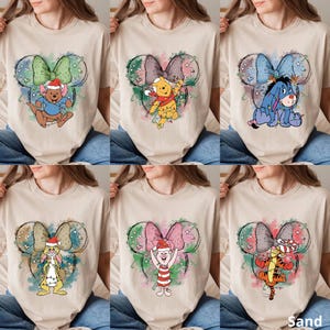 May include: Beige t-shirts featuring a Mickey Mouse ear design with a bow and various Disney characters. Each shirt showcases a different character, including Winnie the Pooh, Eeyore, and Tigger, with festive holiday decorations.