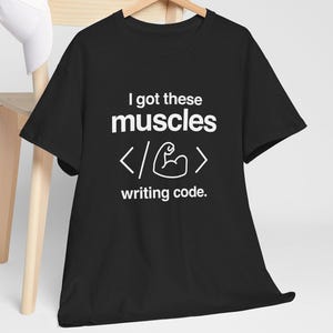 Puede incluir: Camiseta negra con el texto "I got these muscles writing code." El diseño incluye un gráfico de brazo flexionado y símbolos de código. Una gorra de béisbol blanca descansa sobre una silla de madera en el fondo.