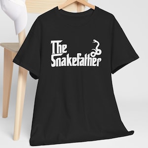 Könnte beinhalten: Schwarzes T-Shirt mit dem weißen Text "The Snakefather" und einer Schlangenillustration. Das Shirt hängt an einem Holzkleiderbügel. Eine weiße Baseballkappe liegt auf einem Holzstuhl im Hintergrund.