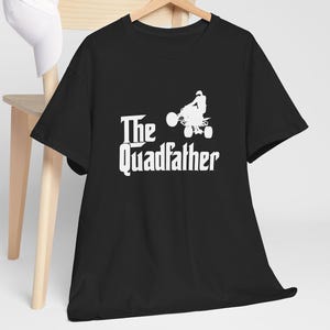 May include: Black t-shirt with the white text "The Quadfather" and a silhouette of a person riding an ATV. The shirt is hanging on a wooden hanger, with a white baseball cap and a wooden chair in the background.