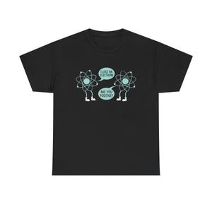 Könnte beinhalten: Schwarzes T-Shirt mit einem Cartoon-Design von zwei Atomen. Eines der Atome sagt: „Ich habe ein Elektron verloren!“, und das andere antwortet: „Bist du positiv?“ Der Text und das Atom-Design sind in Hellblau und Weiß.
