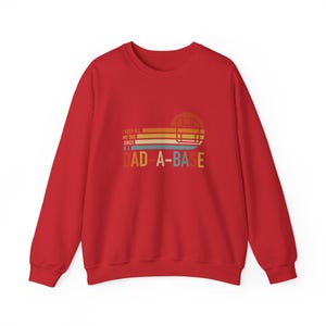 May include: A red crewneck sweatshirt featuring a graphic of a floppy disk inside a sunset circle with horizontal stripes. The text reads: I KEEP ALL MY DAD JOKES IN A DAD-A-BASE. The design uses yellow, orange, and blue colors.