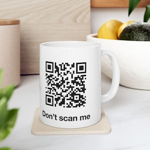 May include: A white ceramic mug featuring a black QR code printed on the front, positioned above the text "Don't scan me" in a simple black sans-serif font. The mug has a standard handle and a smooth, glossy finish.