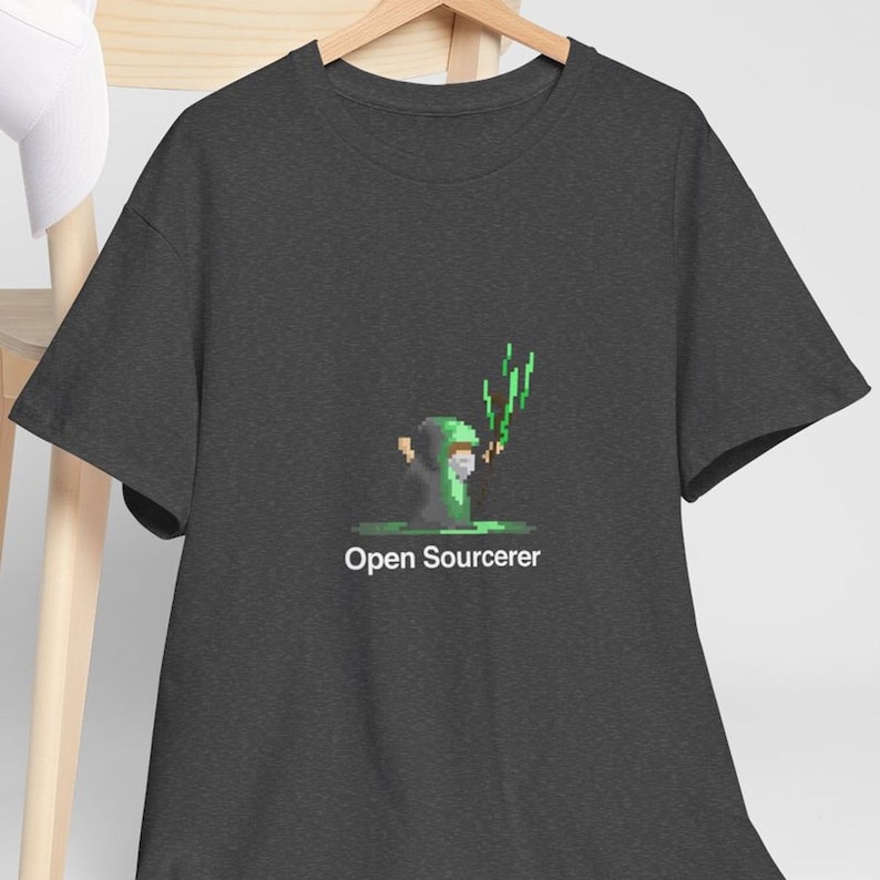 Pu&ograve; includere: T-shirt grigio scuro con una grafica pixelata di un mago con magia verde. Il testo "Open Sourcerer" &egrave; sotto. La maglietta &egrave; appesa a una gruccia di legno.