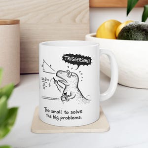 May include: White ceramic mug featuring a cartoon dinosaur wearing glasses and calculating a trigonometry problem. The mug has the text "TRIGGERSOME!" in a speech bubble and "Too small to solve the big problems."