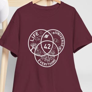 Op de afbeelding: Bordeauxrood T-shirt met een wit grafisch ontwerp. Het ontwerp toont drie overlappende cirkels met de woorden "Life", "Universe" en "Everything". Het nummer "42" staat in het midden van het ontwerp.