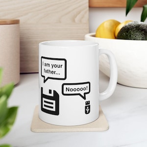 May include: A white ceramic mug featuring a black graphic of a floppy disk with a speech bubble saying I am your father and a small USB drive with a speech bubble saying Nooooo, printed on the front of the cylindrical drinkware.