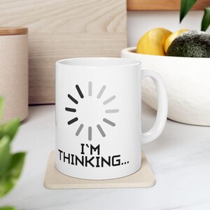 May include: A white ceramic mug featuring a circular loading icon composed of gray and black segments. Below the icon, the text "I'M THINKING..." is printed in a black, pixelated font. The mug has a standard handle and sits on a square coaster.