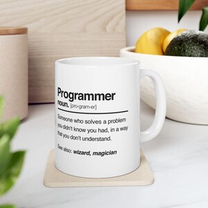 May include: A white ceramic mug featuring black printed text that defines the word Programmer as someone who solves a problem you didn't know you had, in a way that you don't understand, with a See also section listing wizard and magician.