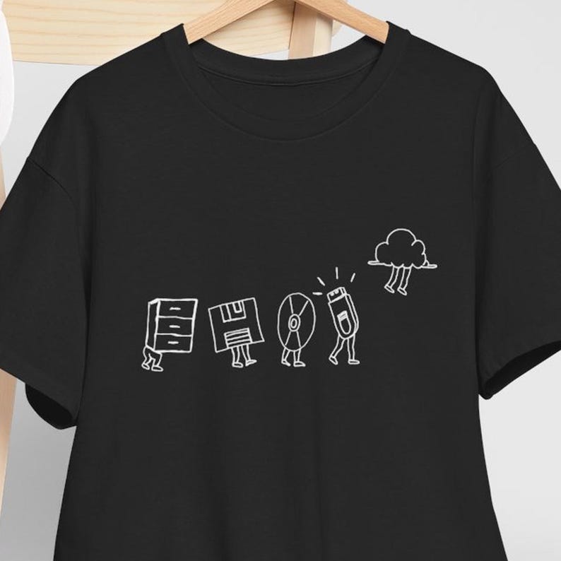 Op de afbeelding: Zwart T-shirt met een witte afbeelding van een diskette, computer, cd, USB-stick en wolk. Het ontwerp is een grillige illustratie van gegevensopslagapparaten. Het T-shirt hangt aan een houten kleerhanger.