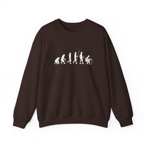 May include: A dark brown long-sleeve sweatshirt featuring a white graphic print on the chest. The design depicts the stages of human evolution, starting with a primate and ending with a figure sitting at a desk working on a laptop computer.