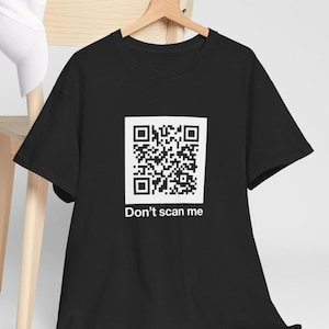 Puede incluir: Camiseta negra con un gran código QR blanco. El texto "Don't scan me" está impreso debajo. La camiseta está colgada en una percha de madera.