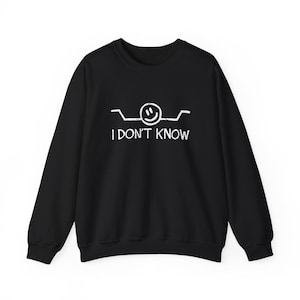 May include: A black long-sleeve sweatshirt featuring a white graphic of a smiling face with shrugging arms and the text I DON'T KNOW printed in a distressed, hand-drawn style font centred on the chest area of the garment.