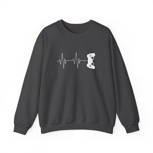 May include: A dark charcoal gray crewneck sweatshirt featuring a white graphic print on the chest. The design displays a horizontal heartbeat line that transitions into the silhouette of a video game controller on the right side.