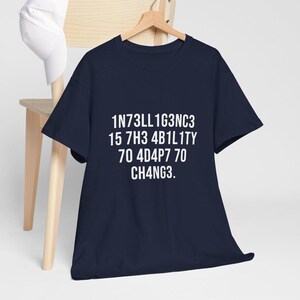 May include: A navy blue t-shirt with white text that reads "1N73LL1G3NC3 15 7H3 4B1L1TY 70 4D4P7 70 CH4NG3." The shirt is hanging on a wooden hanger against a light background.