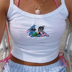 Puede incluir: Camiseta sin mangas blanca con un gr&aacute;fico bordado de dos personajes de dibujos animados con gafas de sol. La camiseta se combina con pantalones cortos a rayas azules y blancas. Un top de bikini de ganchillo rosa y collares de perlas completan el look.