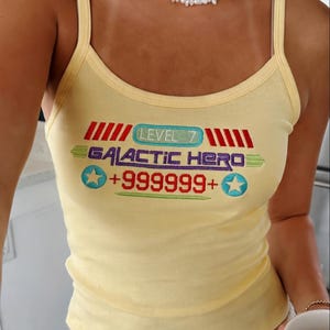 Puede incluir: Camiseta sin mangas de color amarillo claro con tirantes finos. La parte delantera presenta el texto "LEVEL 7 GALACTIC HERO +999999+" en rojo, azul, verde y blanco. Se lleva un collar de perlas alrededor del cuello.