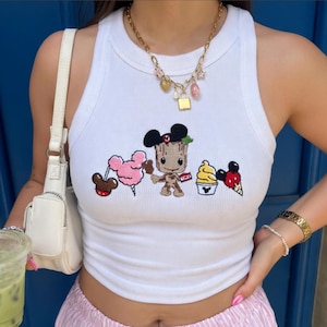 May include: A white ribbed tank top featuring an embroidered graphic of a brown tree-like character wearing black mouse ears, flanked by a pink cotton candy, a yellow soft serve ice cream, and a red ice cream cone with mouse ears.