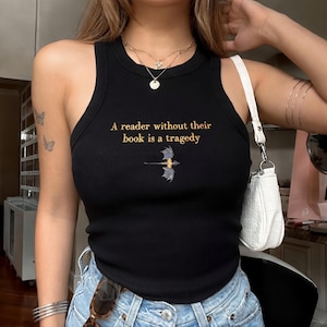 Op de afbeelding: Zwart geribbeld mouwloos shirt met de gouden tekst "A reader without their book is a tragedy". Een kleine drakenafbeelding staat onder de tekst. De top is mouwloos en heeft een ronde hals.