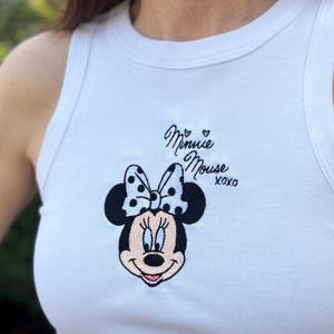 Może przedstawiać: Biały top na ramiączkach z haftowanym wzorem Myszki Minnie. Wz&oacute;r zawiera twarz Myszki Minnie, kokardę w kropki oraz napis "Minnie Mouse XOXO" czarną czcionką. Swobodny top bez rękaw&oacute;w.