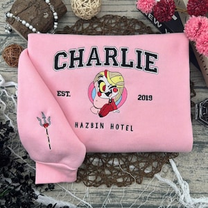 May include: A light pink sweatshirt with the name "CHARLIE" embroidered in black block letters. Below is a cartoon character and the words "EST. 2019 HAZBIN HOTEL". The left sleeve has a trident design.