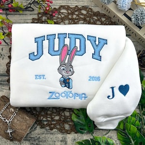 Puede incluir: Sudadera blanca con texto bordado en azul "JUDY" y un personaje de conejo de dibujos animados. La sudadera tambi&eacute;n presenta el texto "EST. 2016" y "ZOOTOPIA". La manga tiene un coraz&oacute;n azul y la letra "J".