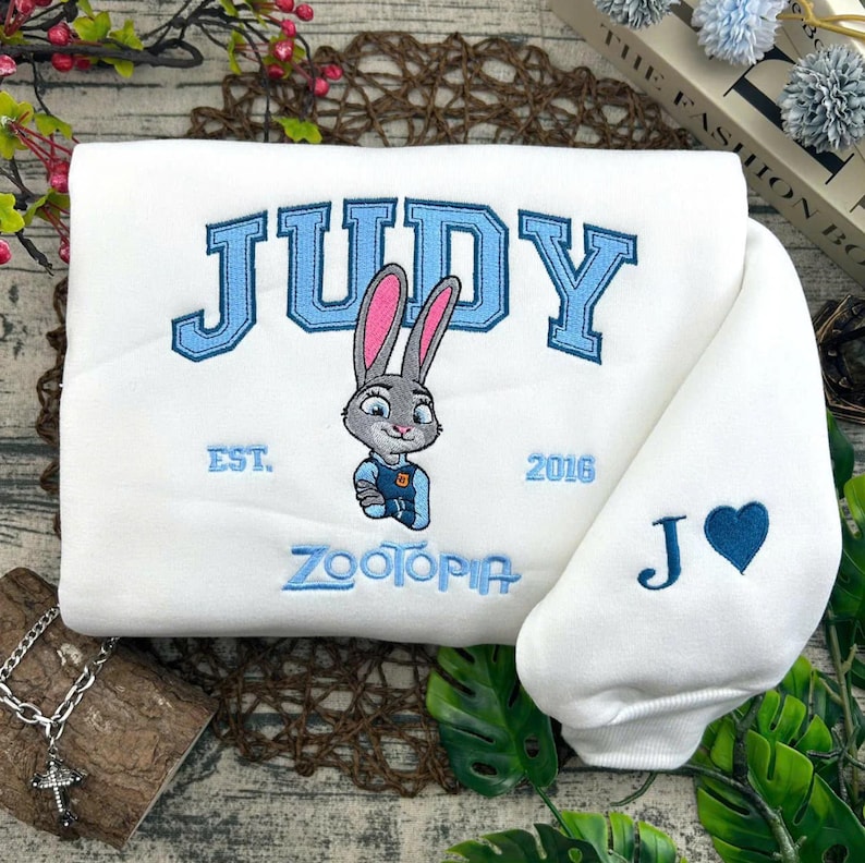 Puede incluir: Sudadera blanca con texto bordado en azul "JUDY" y un personaje de conejo de dibujos animados. La sudadera tambi&eacute;n presenta el texto "EST. 2016" y "ZOOTOPIA". La manga tiene un coraz&oacute;n azul y la letra "J".
