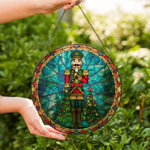 May include: A round stained glass window featuring a nutcracker soldier in a green and red uniform. The soldier stands between two green Christmas trees. The window has a blue and green background with a decorative border and is hanging from a chain.